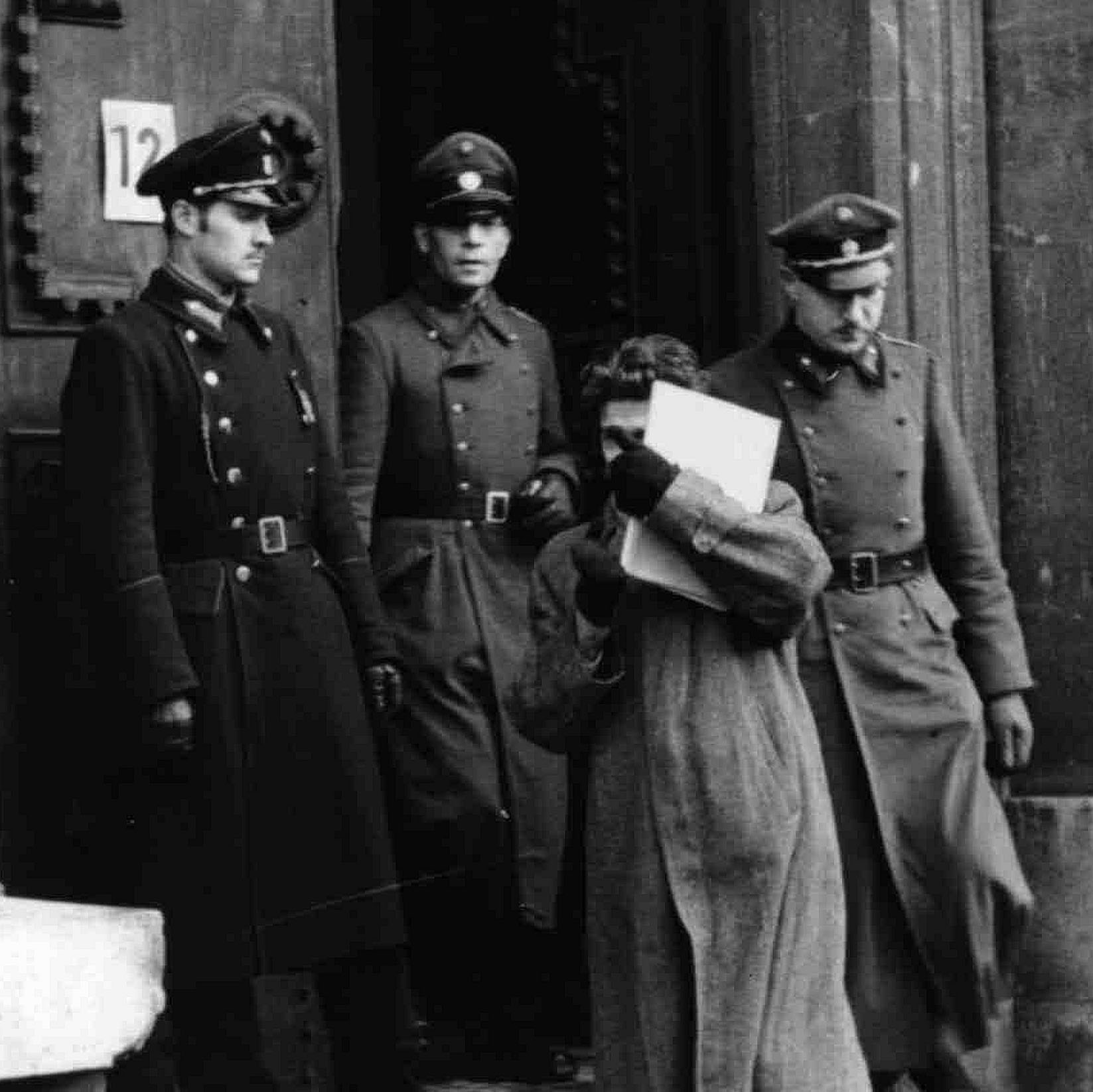 Johann Pav on the stairs to the courthouse, his face hidden behind a file. Three uniformed men are standing behind him.